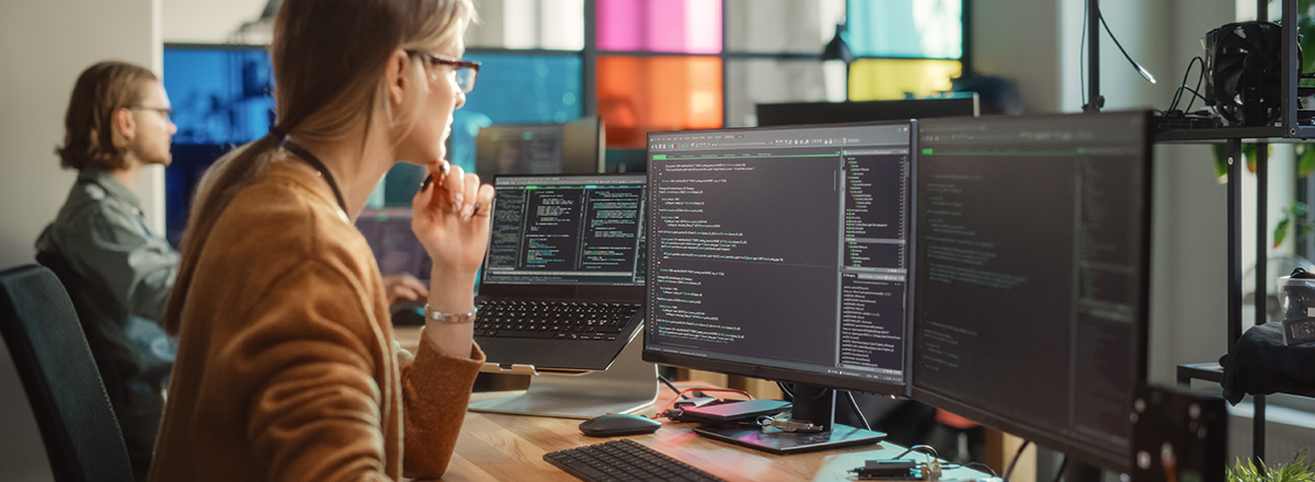 A woman in an office looking at code on multiple computer monitors.