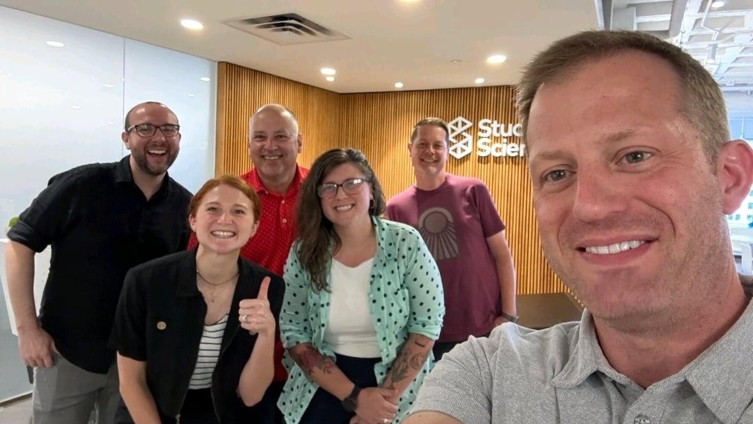 Steve Pruden takes a selfie in the Studio Science headquarters lobby with several team members smiling behind him.
