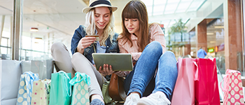 Two women shopping on an iPad surrounded by shopping bags.